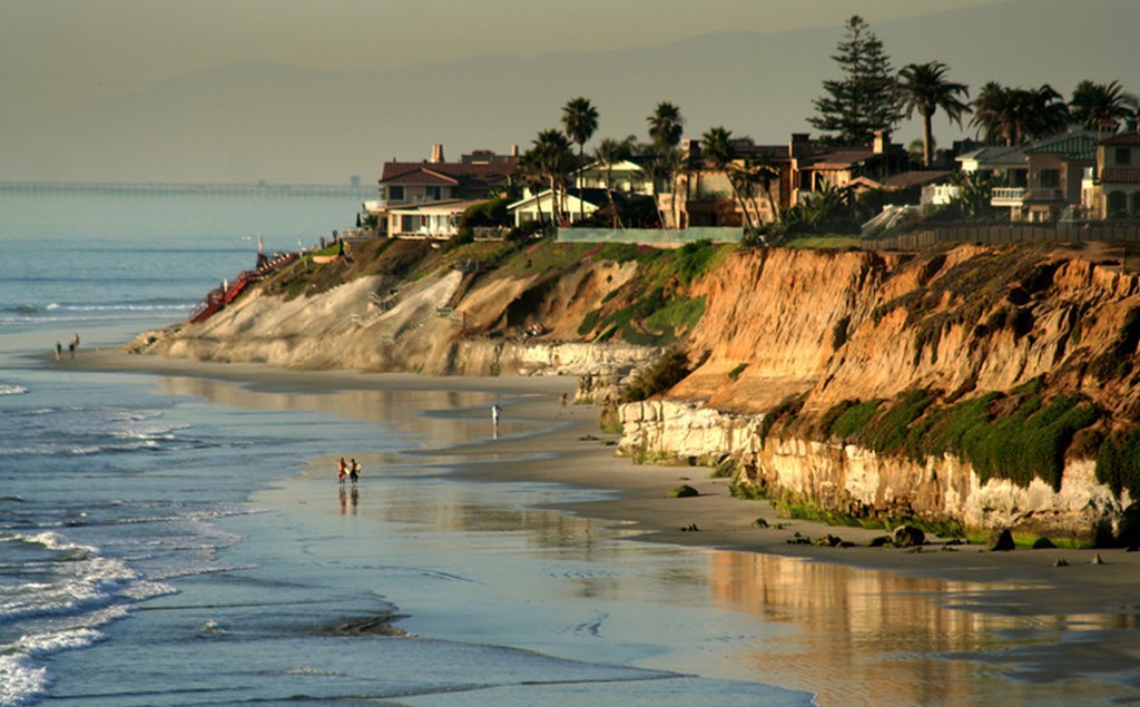 Coastal houses atop cliffs overlooking a sandy beach with ocean waves and distant mountains.