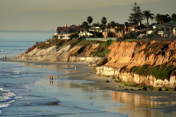 Coastal houses atop cliffs overlooking a sandy beach with ocean waves and distant mountains.