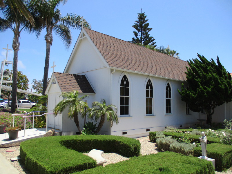 Small white chapel with brown roof, surrounded by palm trees and shrubs.