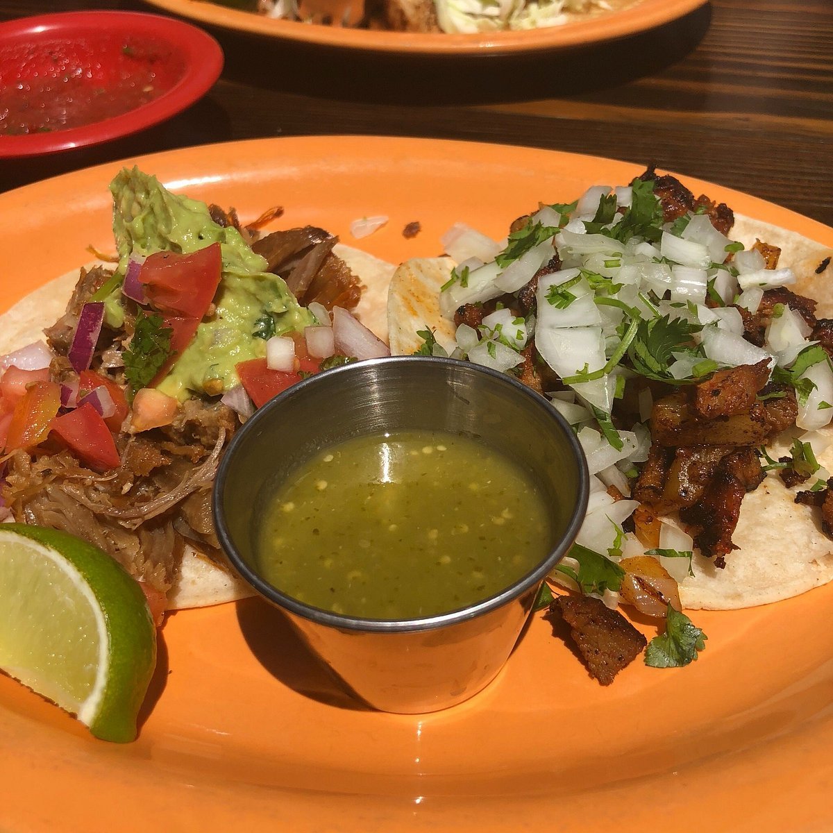 Two tacos with beef and pork, garnished with guacamole, onion, cilantro, and lime, served with green salsa.