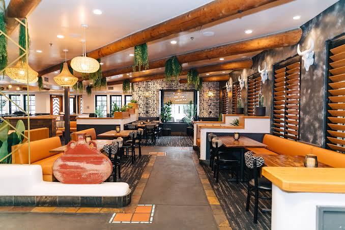 Rustic restaurant interior with wooden beams, orange booths, and decorative plants.