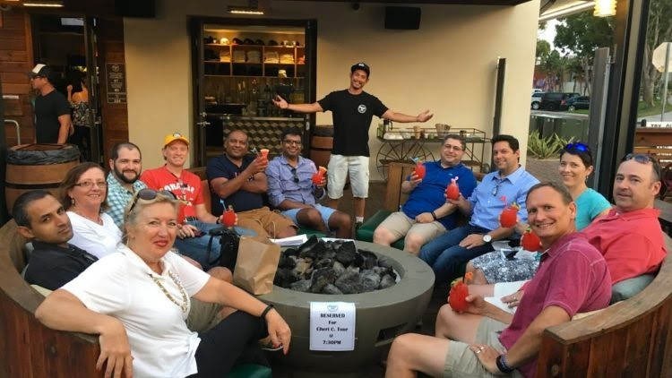 Group of people sitting around a fire pit holding red drinks, outdoors at a restaurant.