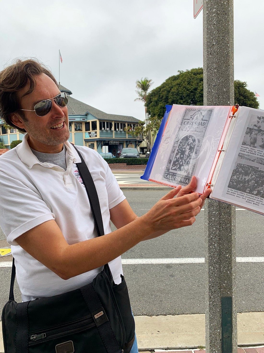 Man in sunglasses holding a binder on a street, near a pole with shops in the background.