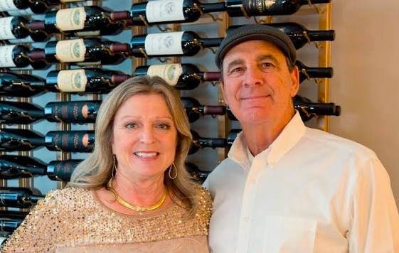 Two people smiling in front of a wall-mounted wine rack.